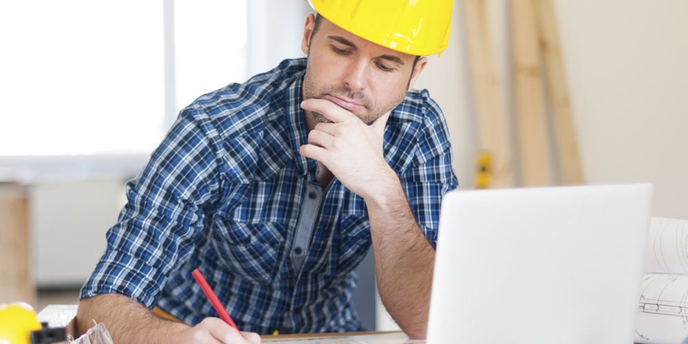 Man in yellow hardhat working on laptop and building plans