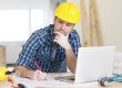 Man in yellow hardhat working on laptop and building plans