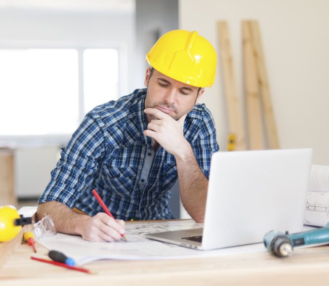 Man in yellow hardhat working on laptop and building plans